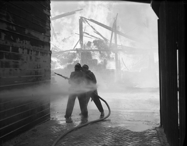 821061 Afbeelding van twee brandweerlieden tijdens het blussen van een brand bij Houthandel Jongeneel (Zeedijk 6) te Utrecht.